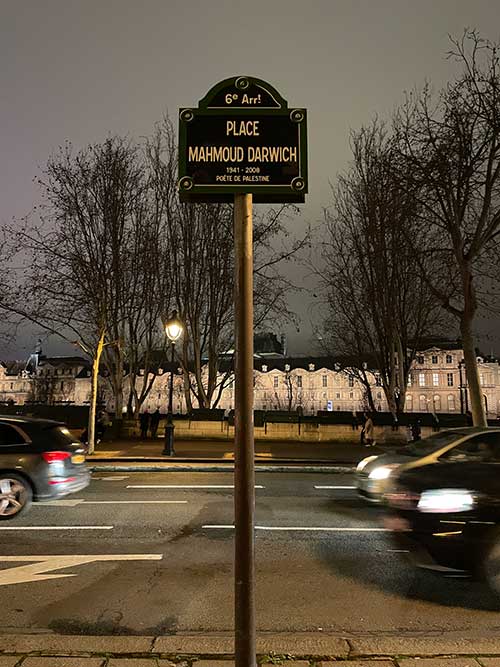 A photograph of a street sign in Paris. It reads: “6th Arrondissement/Mahmoud Darwish Square/1941–2008/Poet of Palestine.” Behind the sign is the Louvre Museum, situated on the other side of the Seine River.