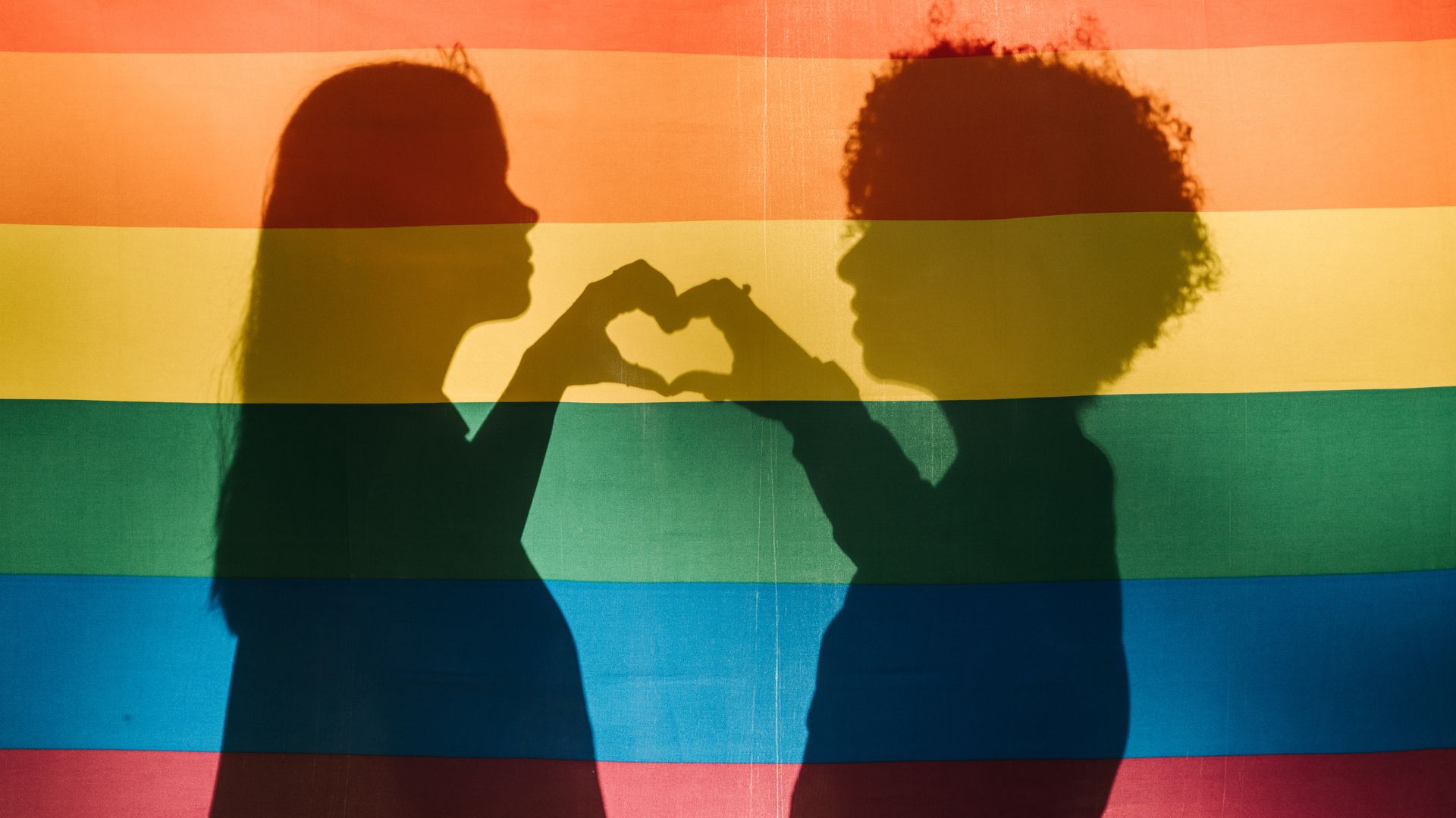 Two silhouettes of women behind a pride flag making a heart together with one hand from each coming together