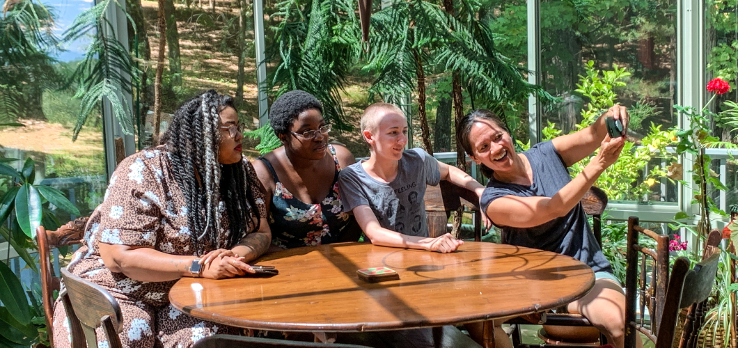 Four writers sit on a table in a sunroom under the sun. There are trees outside.