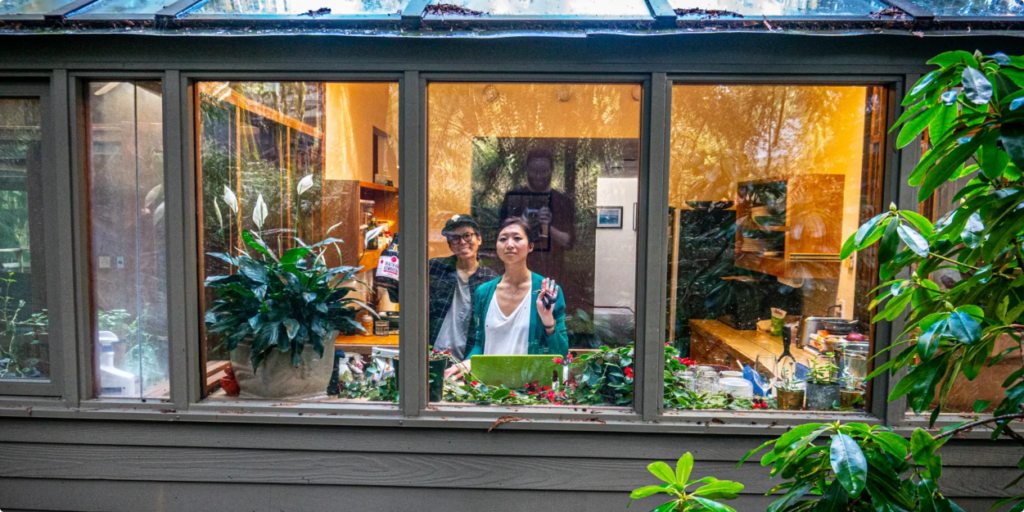 two individuals waving at the camera through a window. They are in the kitchen of a house. The reflection of the photographer is visible on the window's surface