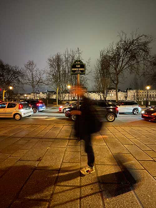 A photograph of a street sign in Paris. It reads: “6th Arrondissement/Mahmoud Darwish Square/1941–2008/Poet of Palestine.” There is a  person pulling a suitcase and walking past it quickly.